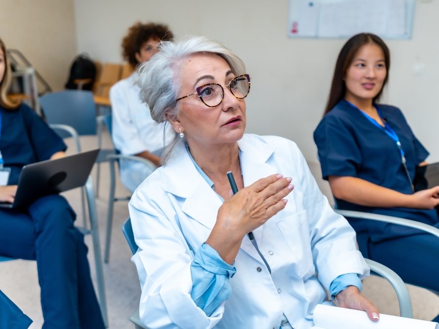 Clinical staff in scrubs and white coats seated in a training session, one participant speaking while others take notes