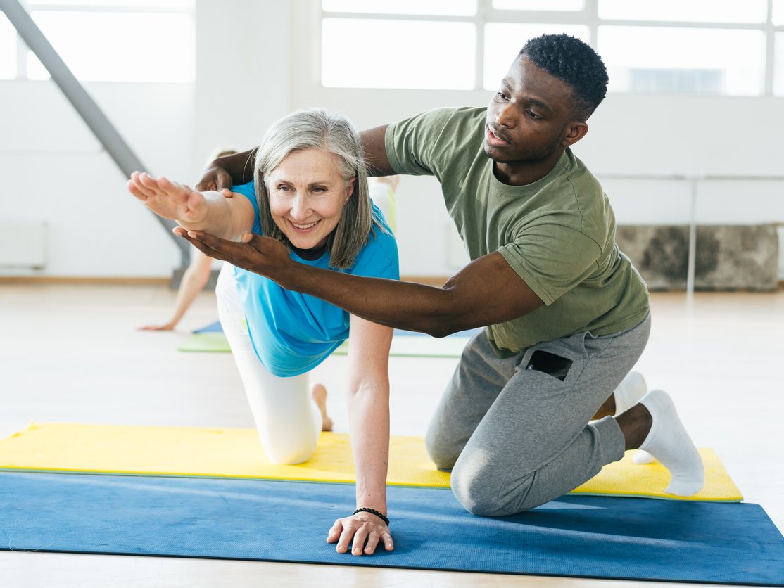 A specialist supporting an older woman through a quadruped balance position on a mat, hand guiding her outstretched arm